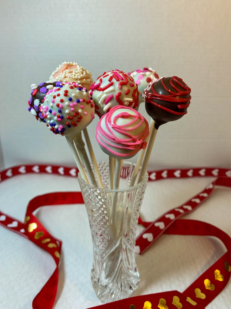 White and milk chocolate truffles decorated in pink, red and purple decorations to celebrate Valentine's Day, displayed in a decorative glass with red ribbons